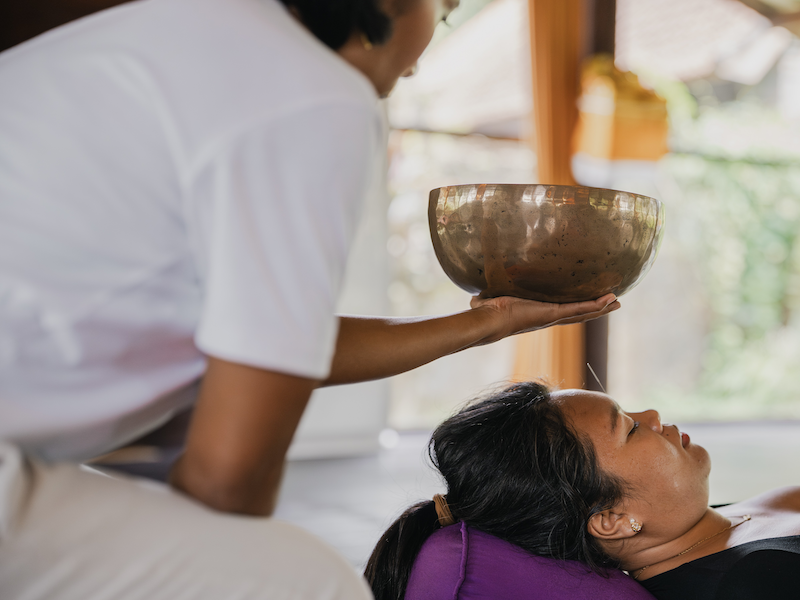 A practitioner holds a Tibetan singing bowl above a resting participant receiving acupuncture
